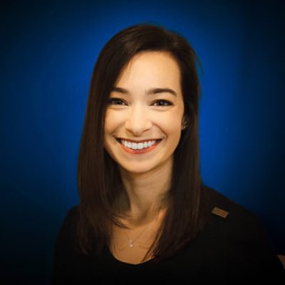 The image shows a woman with short hair smiling at the camera. She has dark hair and is wearing a black top. Behind her, there s a blue background that appears to be a digital wallpaper.