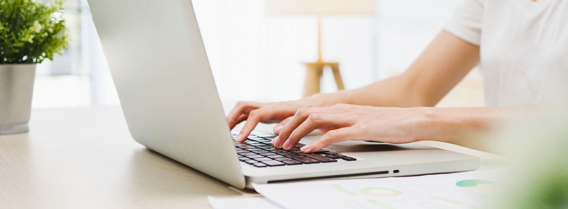 The image shows a person using a laptop computer on a wooden desk with a blurred background featuring indoor plants and a portion of another person's upper body, suggesting a collaborative workspace environment.