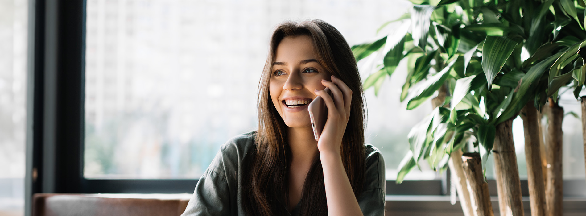 A woman sitting at a table with a plant behind her, smiling while talking on her cell phone.