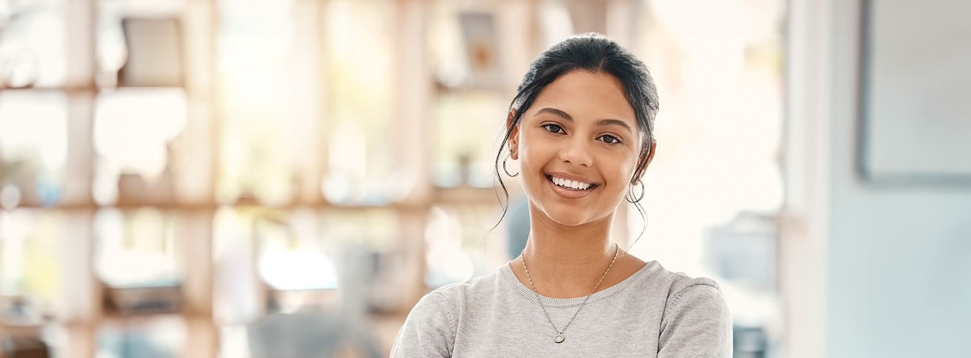 The image shows a young woman with a smile, standing inside a library-like setting, wearing a light grey top.