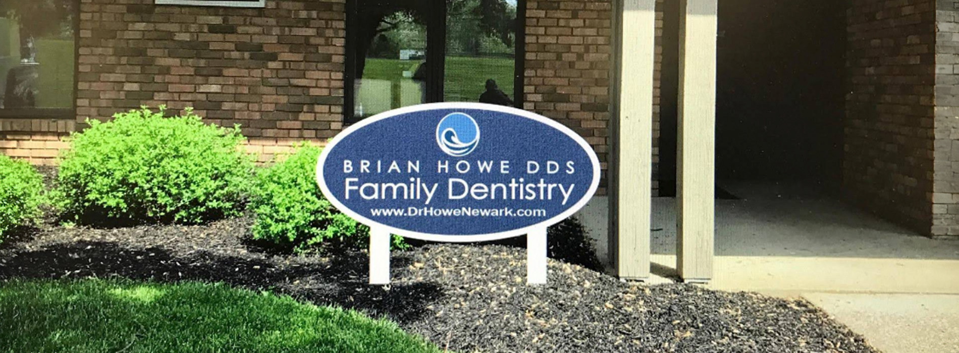 The image shows a sign for Brian Howe DDS Family Dentistry with the name Brian Howe displayed on it, placed in front of a building with a brick facade, surrounded by landscaping and trees.