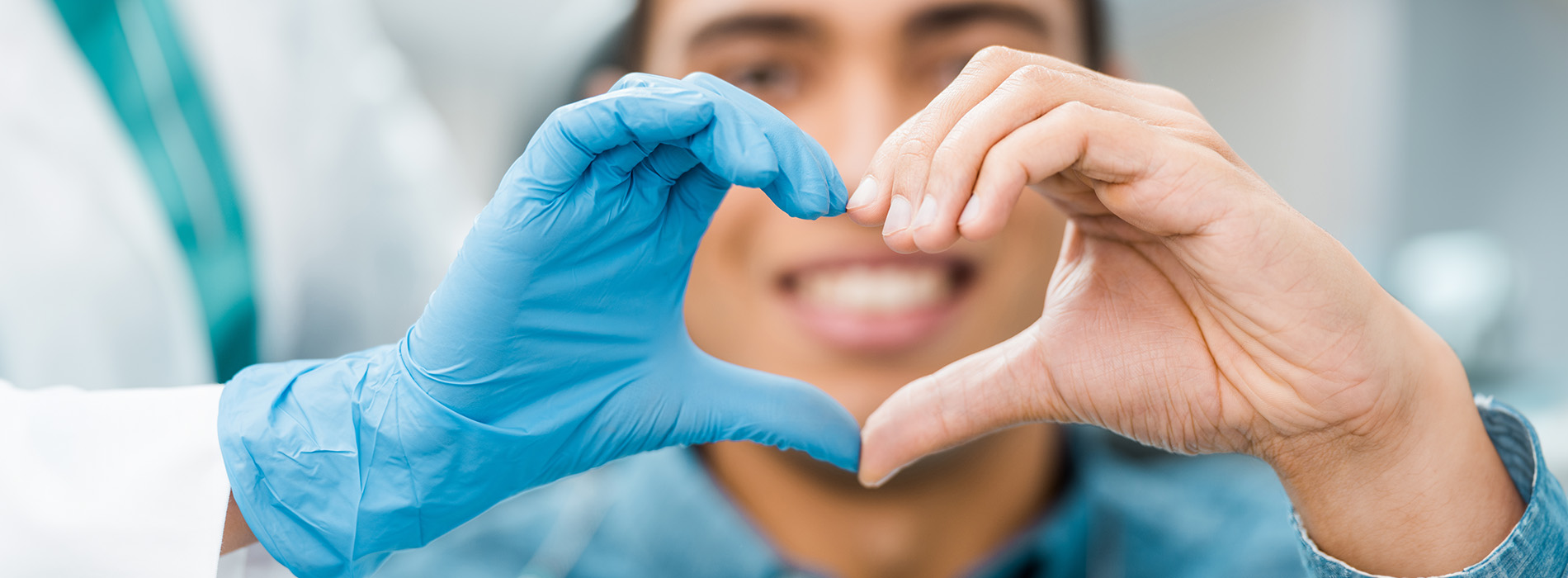 The image shows two hands forming a heart shape against a background featuring a person wearing blue gloves and a medical setting.