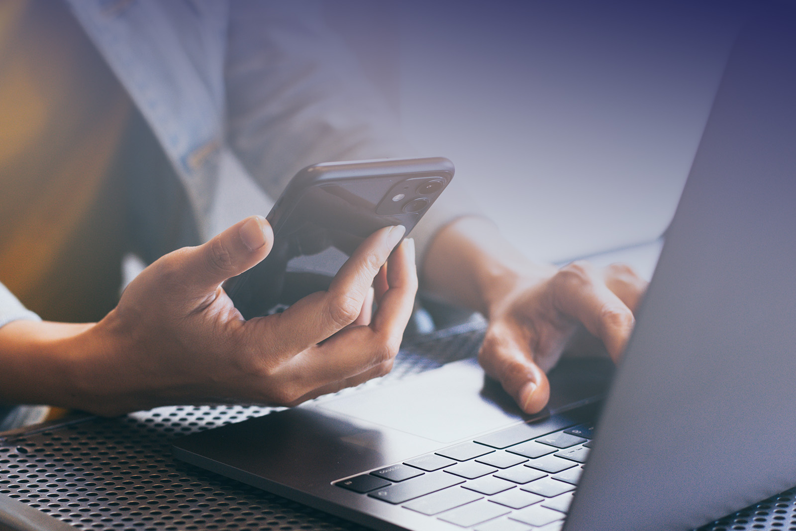 The image shows a person's hands holding a smartphone while sitting at a table with a laptop open in front of them.