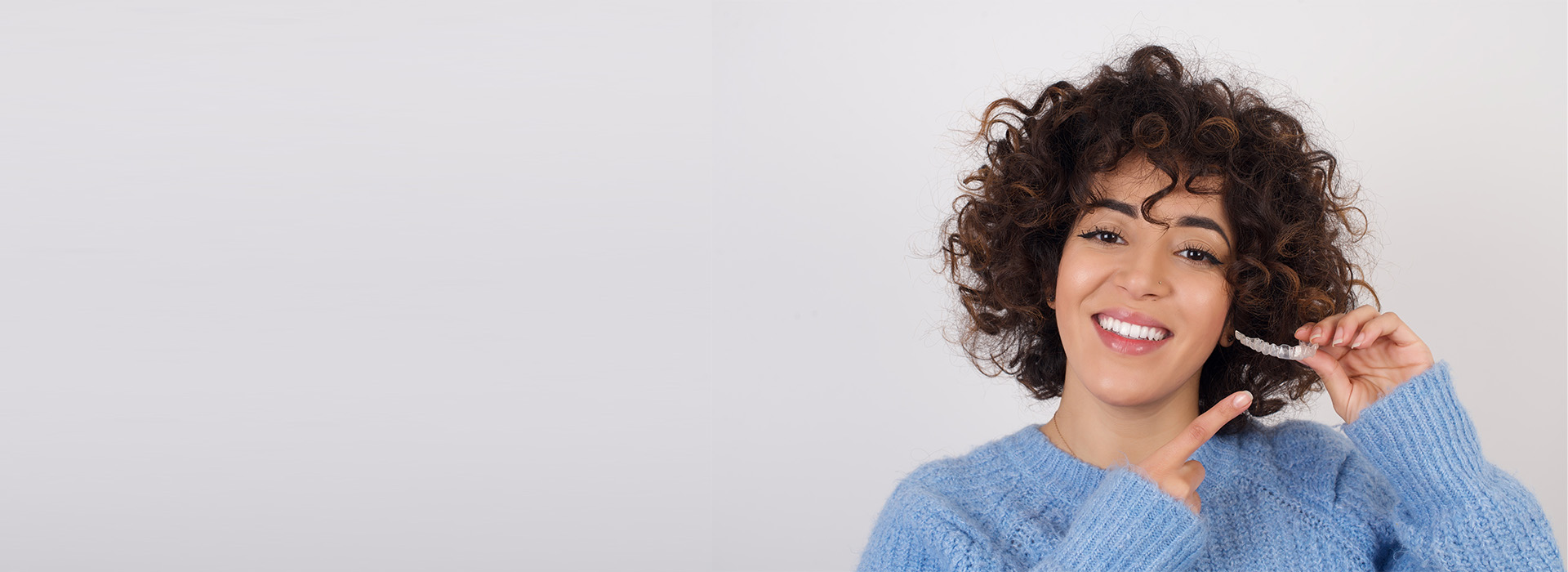 A woman with curly hair brushing her teeth smiles at the camera while holding a toothbrush.