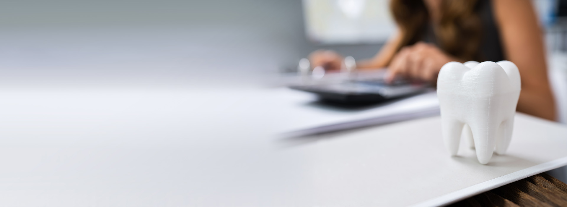 The image shows a person working at a desk with a computer monitor, surrounded by papers, while a small white figurine of a tooth is placed on the desk, possibly indicating dental care or a dental-related business context.