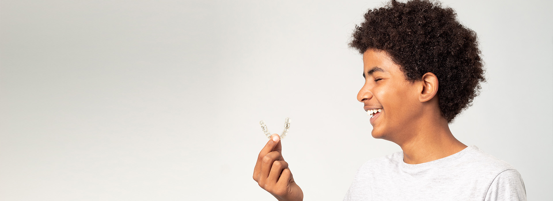 The image shows a person with an afro hairstyle smiling broadly while holding a small object between their fingers, set against a plain background.