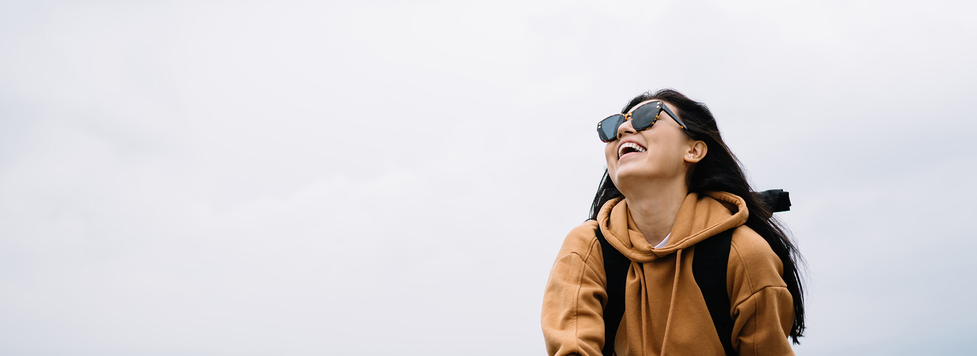 The image shows a person from behind, wearing a backpack and sunglasses, standing on a cloudy day with a clear sky visible in the background.