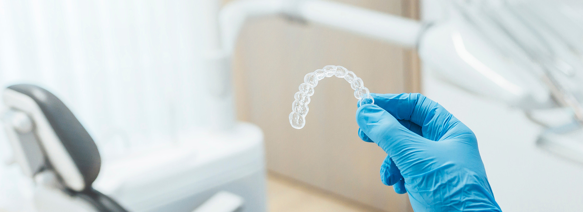 A hand holding a transparent glove with an object inside it, placed against a blurred background featuring dental equipment.
