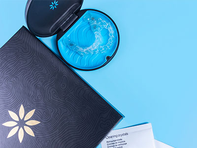 The image shows a blue box with a gold emblem on its side placed next to an open blue container with a clear lid containing a green substance, possibly a skincare product, against a teal background.