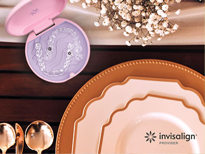 The image features a table setting with various dishes, silverware, and decorative items including a pink toothbrush holder with a design on it, placed on a wooden surface with a backdrop of a white wall and a floral arrangement.