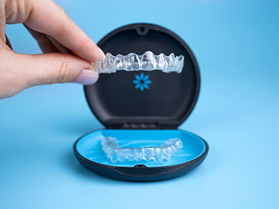 A hand holding a clear plastic tray containing a set of transparent dental aligners with a blue case in the background.