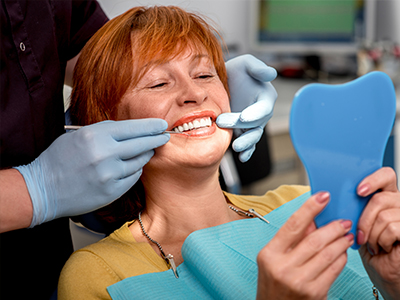 The image shows a woman sitting in a dental chair with a blue tooth-shaped object held up to her face, smiling at the camera while being attended to by dental professionals who are adjusting her mouthpiece.