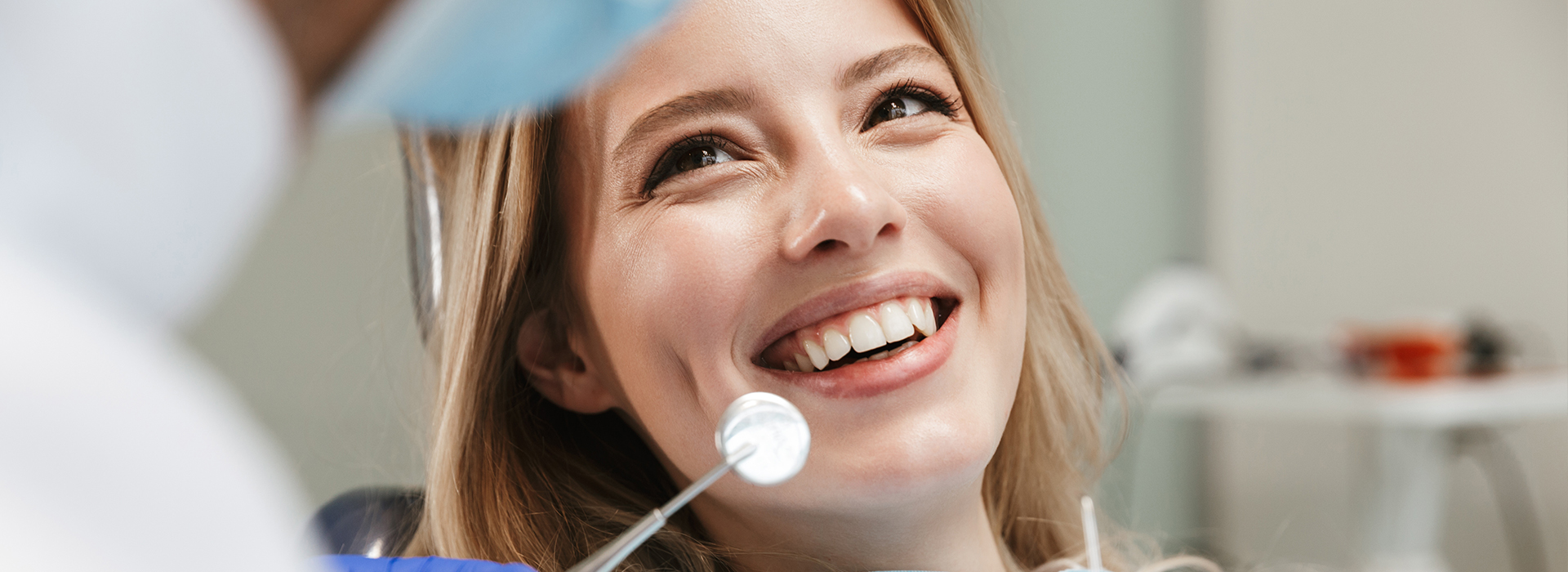 The image shows a smiling woman sitting in a dental chair with a dentist s mirror in front of her, undergoing a dental appointment.