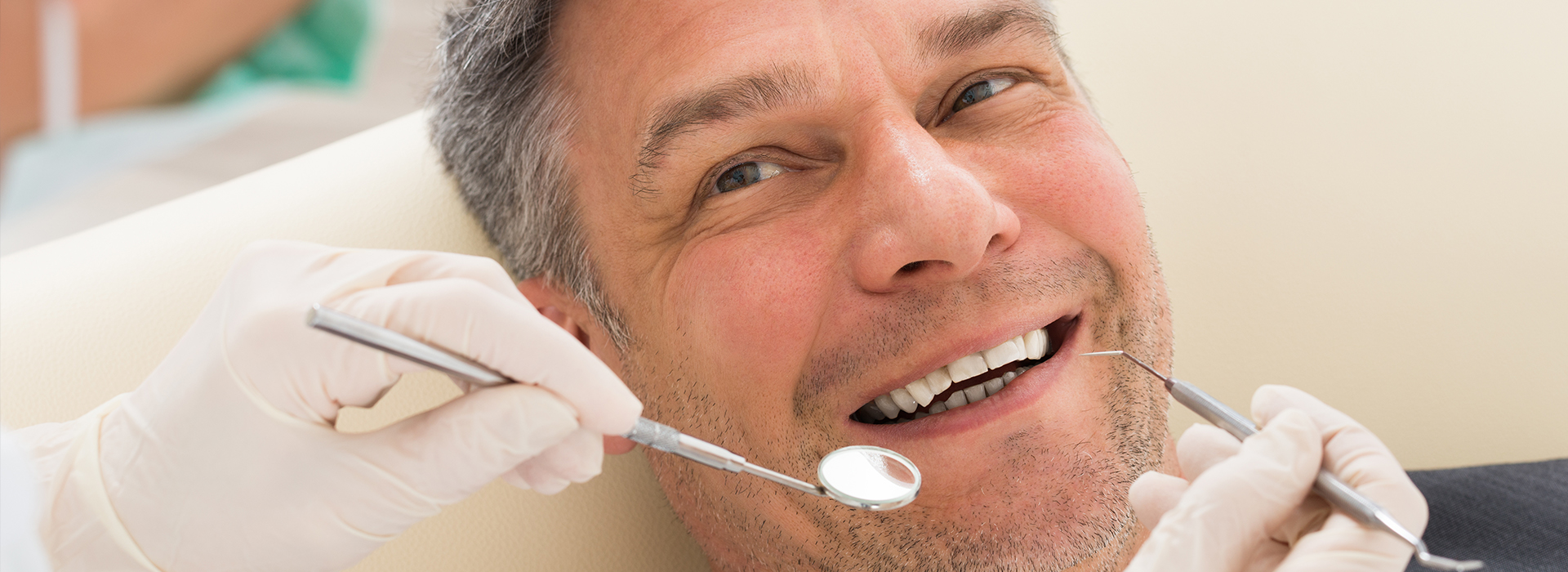 The image shows a man sitting in a dental chair with his mouth open, receiving dental treatment, with visible dental instruments such as a scaler being used by a dental professional out of frame.