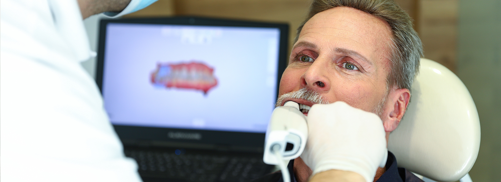 The image shows a man receiving dental care, with a medical professional assisting him while he is seated in a dental chair.