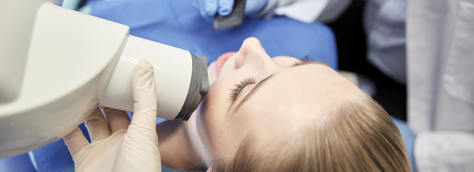 This image depicts a person receiving a medical treatment, likely at a dental clinic, with a magnified view of their mouth through a device resembling a microscope, while another individual appears to be operating the equipment.