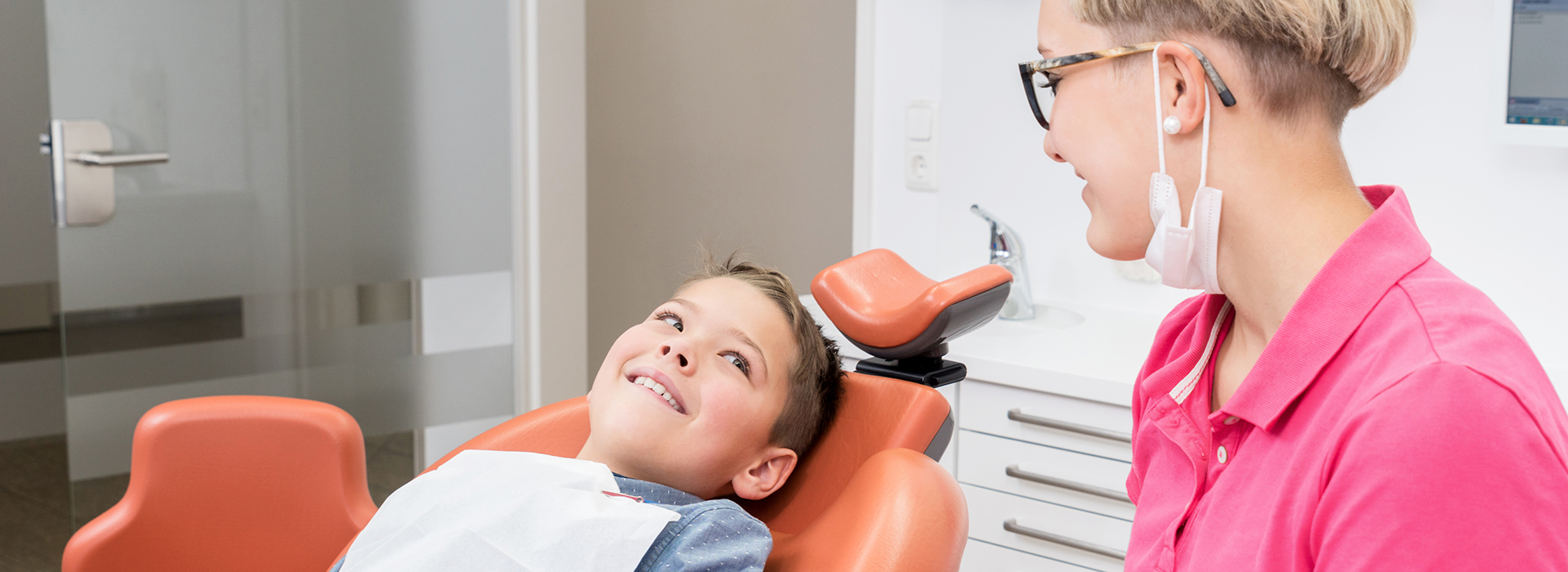 The image shows a person sitting in a dental chair with their head turned towards a woman who appears to be a dental professional, possibly during a dental examination or cleaning procedure.
