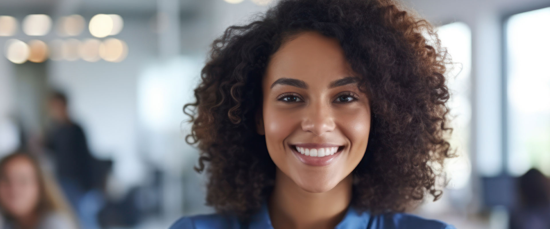 The image shows a smiling woman with curly hair, wearing a blue top, standing in an indoor setting that appears to be a professional office environment.
