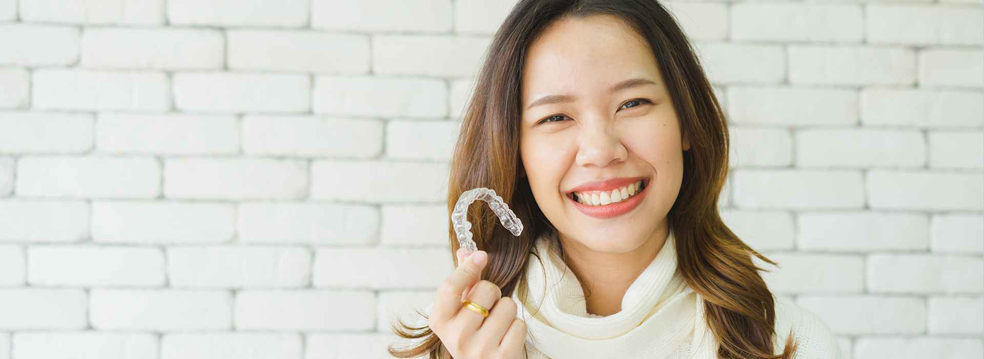 A young woman with a bright smile holds up a toothbrush while standing against a brick wall background.
