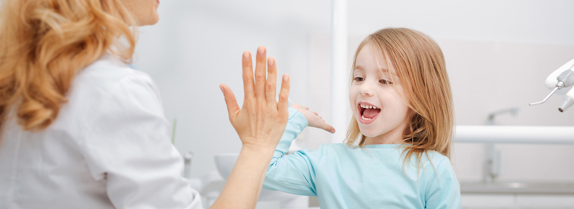 A child with blonde hair, wearing a blue shirt, reaches up towards an adult s hands, which are raised near their face in a gesture of high-five or greeting. The adult appears to be a woman with brown hair, smiling and looking down at the child. They are both indoors, possibly in a bathroom given the presence of a sink in the background.