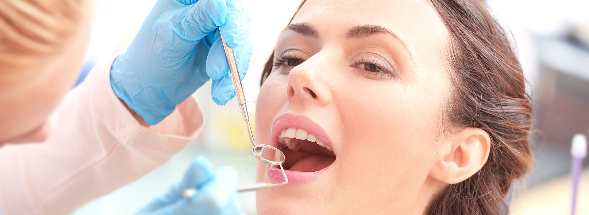 The image shows a woman receiving dental care, with a dentist performing an examination using a mirror and dental instruments while the patient looks on attentively.