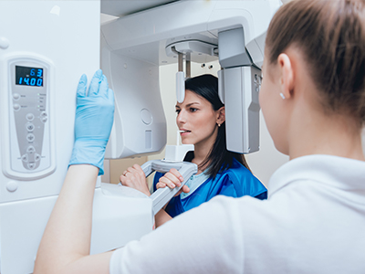A woman in a blue coat is standing next to a large 3D scanner machine, with another woman in a white coat looking at her through the scanner s screen. Both women are wearing face masks and gloves, indicating a health-related setting.