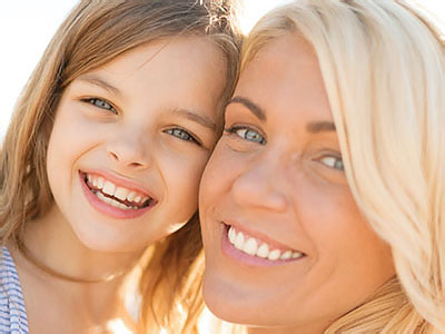 The image shows a woman with blonde hair smiling at the camera, standing next to a young girl who also smiles at the camera. Both are outdoors under clear skies.