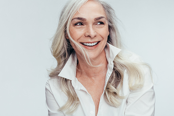 The image shows a woman with blonde hair, wearing a white shirt, smiling and looking slightly away from the camera.