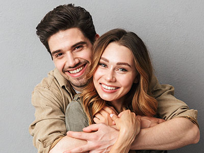 The image shows a man and a woman hugging each other with smiles on their faces  they are both smiling at the camera, and the man has his arms wrapped around the woman from behind.