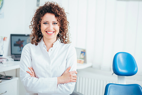 The image shows a woman standing in an office setting with a professional appearance, smiling at the camera.
