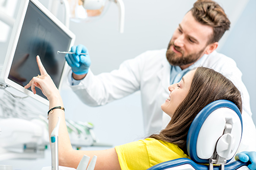 This is a photograph showing an adult woman seated in a dental chair with a dental professional standing beside her, both in a modern dental facility.