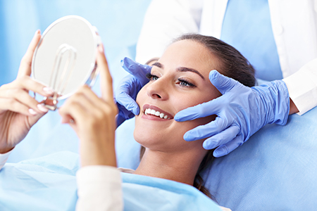 The image depicts a woman lying on a dental chair with her eyes closed, receiving dental treatment under the supervision of a dentist who is using a magnifying glass and wearing gloves.