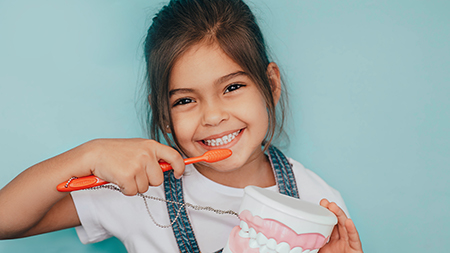 A young girl brushing her teeth with an electric toothbrush, holding a tube of toothpaste.