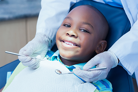 The image shows a young boy sitting in a dental chair, smiling at the camera while receiving dental care, with a dental professional wearing gloves and a mask attending to him.