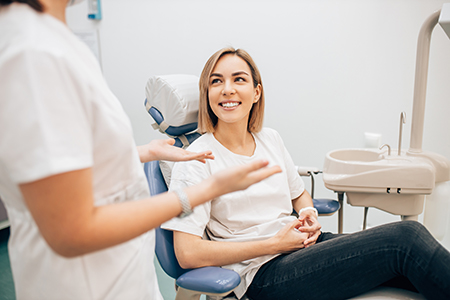 A woman sits in a dental chair, smiling at someone off-camera, while a dentist attends to her with a tray of dental instruments nearby.