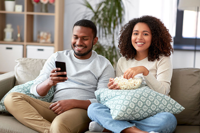 The image shows a man and woman sitting on a couch, with the man holding a smartphone, both looking at the screen. They are engaged in an activity together, likely watching something on the phone or sharing content.