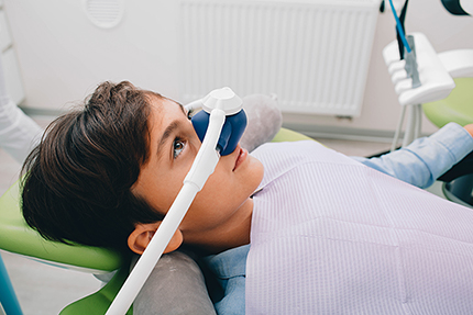 A young person sitting in a dental chair with an examination device over their eyes, wearing a blue surgical mask, and looking at the camera.