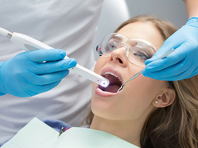 The image shows a person receiving dental care with a device that appears to be an impression-taking tool being used by a dental professional, while another individual monitors the process closely using a digital device.