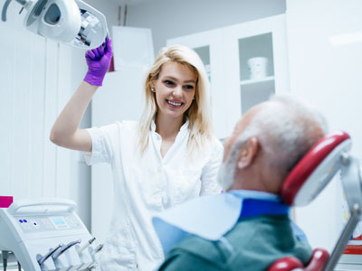 A woman in a white coat smiles as she holds a dental device near an elderly man s face, who is seated in a dental chair with his eyes closed.