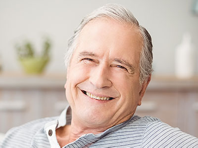 The image shows a smiling older man with gray hair, wearing a blue shirt, sitting in a relaxed posture with his arms crossed on a couch.